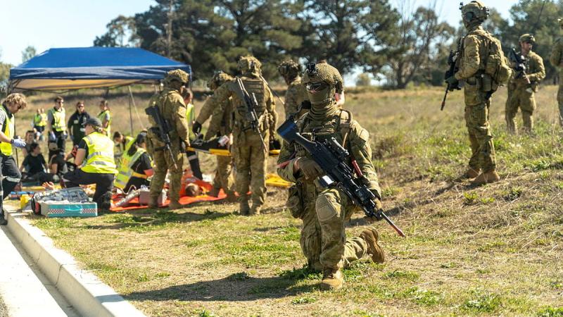 A soldier in camouflage kneels on grass with a rifle while several people stand behind near a small gazebo.