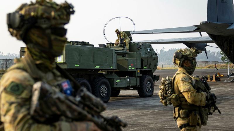 People in uniform, with their faces covered, hold weapons on an airfield tarmac while a large military vehicle sits near the hold of a large military aircraft.