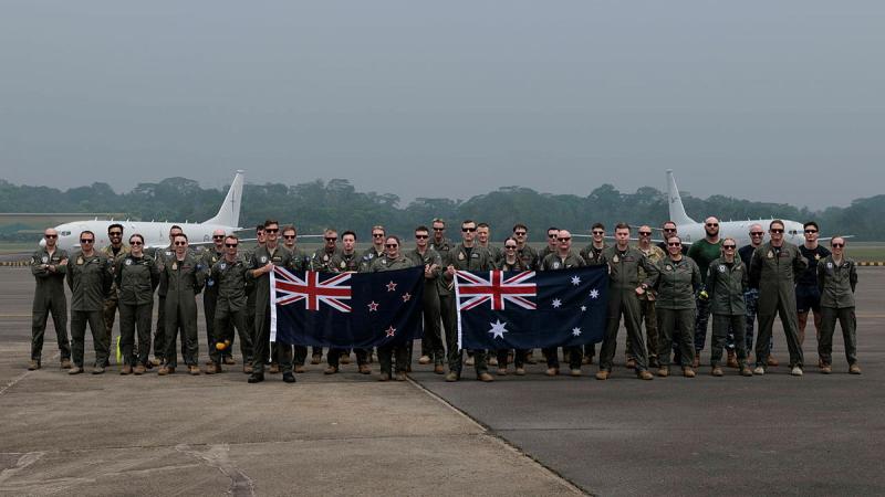 A group of military personnel, holding a NZ flag and an Australian flag, stand in front of two aircraft.