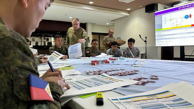People in uniform sit around a large table looking at sheets of paper, while a large projector screen shows a slide in front of the table.