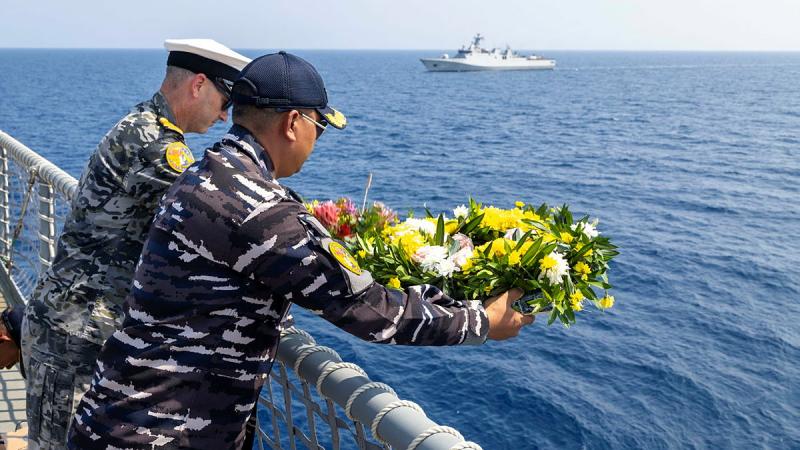 Two men in uniform look over the rail of a ship while one holds a wreath above the water.