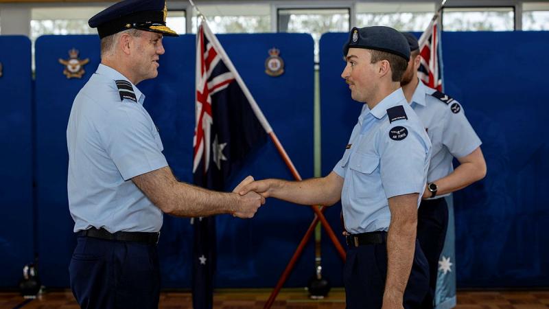 Two male military members shake hands in front of an Australian flag.