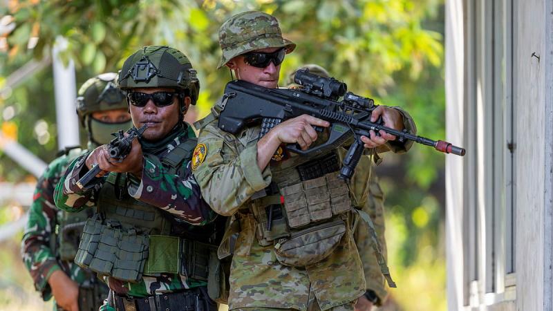 Military personnel walk through a street with guns at the ready.