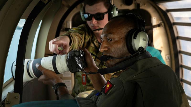 A military man instructs another military man with a camera, while both are inside an aircraft.