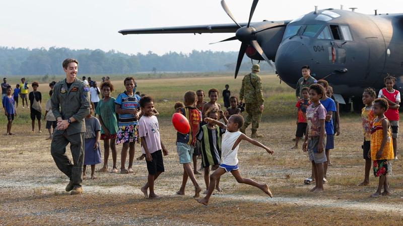 A military man watches a child kick a football. Other children and an aircraft are nearby.