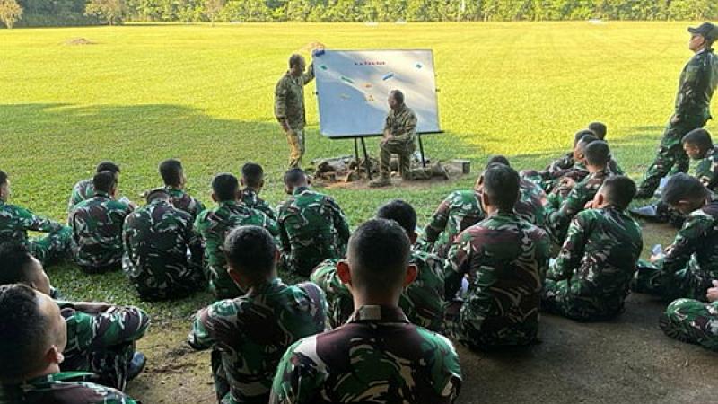 People in uniform sit on the ground outdoors while looking at two people standing at a whiteboard in front of them.