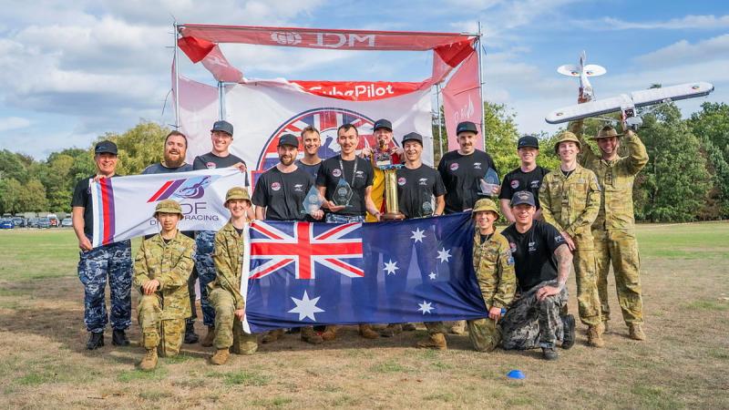 A group of military personnel stand with an Australian flag, a trophy, a drone and a drone racing banner. 
