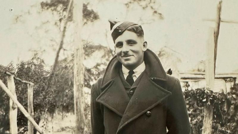 A young man wearing a coat and uniform cap stands outdoors in a black-and-white photograph.