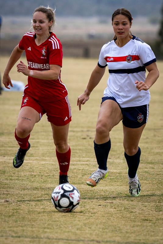 Two female football players from opposite teams chase a soccer ball on a grassy field.