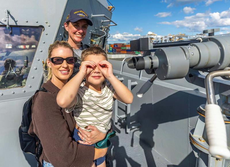 A Navy member, standing near large fixed binoculars on board a ship, stands with a mother and her son who is making the shape of binoculars against his eyes with his hands.