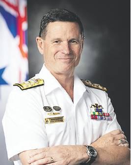 A person wearing a white naval dress uniform with shoulder insignia, ribbons, medals, and qualification badges poses with folded arms. A flag is partially visible in the background, and the portrait is set against a dark studio-style backdrop.