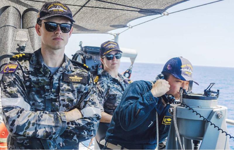 Three naval personnel in camouflage and dark-blue uniforms stand on the deck of a ship at sea beneath a shaded canopy. In the foreground, one person wearing sunglasses and a cap stands with arms crossed. Behind them, a second person in similar uniform and sunglasses stands near ship equipment. On the right, a third person uses a mounted optical navigation instrument while holding a communication handset. The ocean and horizon are visible in the background. Visible text includes the name “REES” on a uniform 
