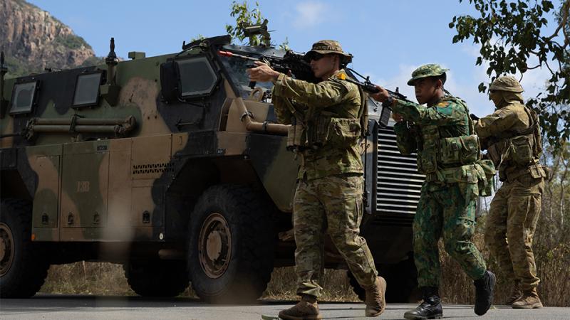 Three people in camouflage uniform walk in a line with weapons raised past an armoured personnel carrier.
