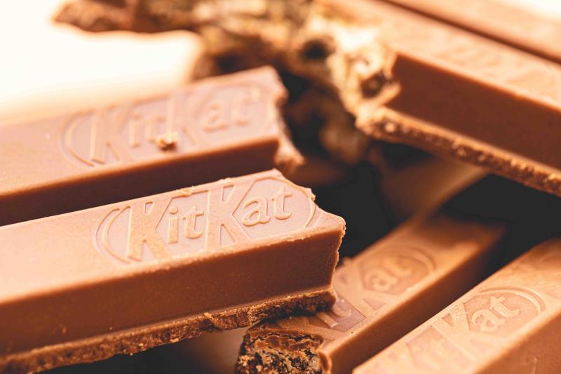 Close-up of several KitKat chocolate wafer bars stacked and overlapping, showing the embossed KitKat logo on the milk chocolate surface and the layered wafer filling inside broken pieces.