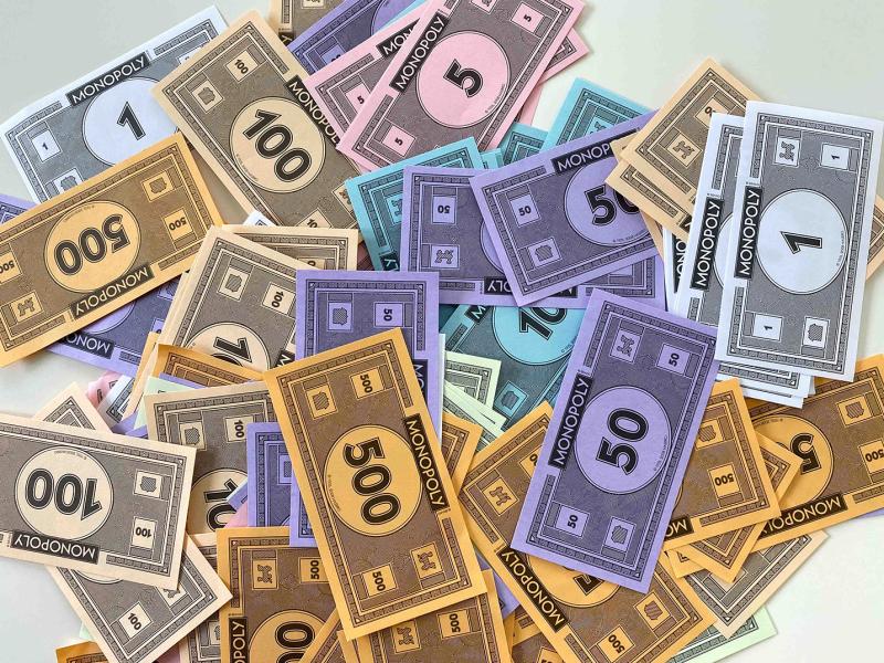 A pile of colourful Monopoly play money notes in various denominations, including 1, 5, 10, 50, 100 and 500, scattered across a light background.