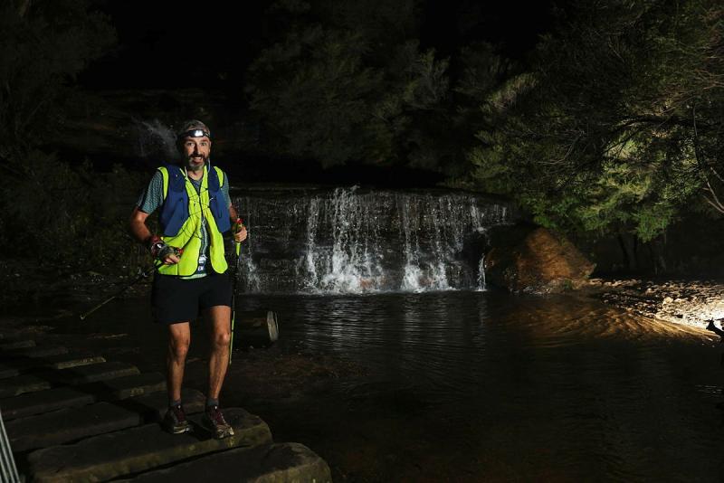 A man in high-vis hikes at dark past a small waterfall.
