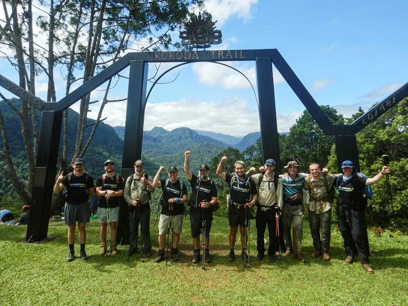 A group of people stand beneath the sign for the Kokoda Trail, with a mountain range in the background.