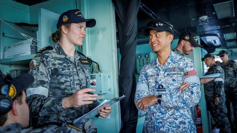 A Navy sailor holding a clipboard speaks to people while on a ship.