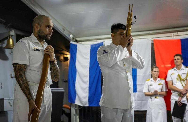A sailor blows into a Japanese instrument while another sailor stands beside him with a didgeridoo.