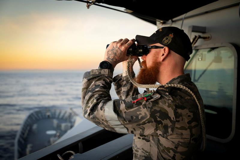 A sailor looks out to sea from a ship, through binoculars.