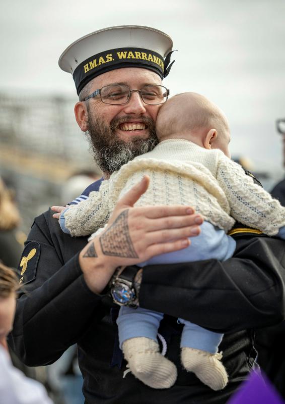 A sailor with a beard and glasses holds and hugs his child, wearing a pale yellow knitted jumper.. 