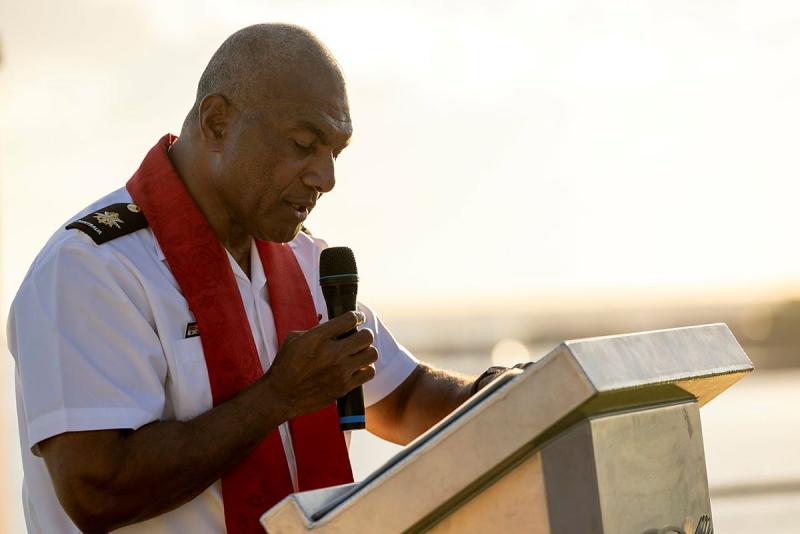 A chaplain speaks into a microphone against a sunset backdrop.