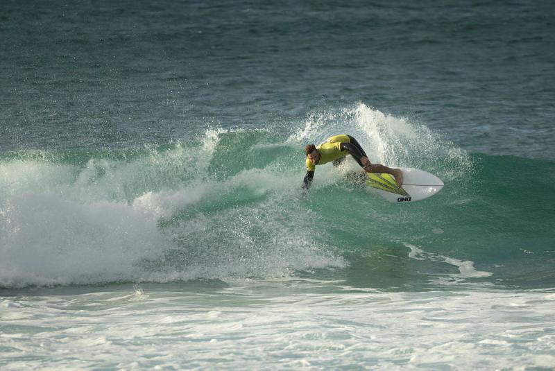 A Navy surfer wearing a yellow jersey and black spring suit carves up a wave.