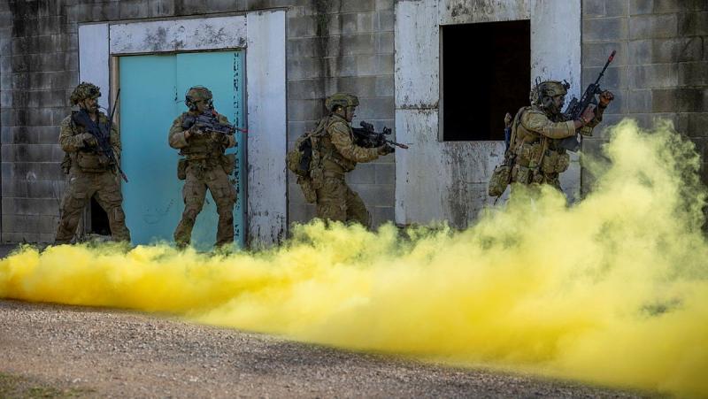 Soldiers with guns run close to a building as yellow smoke fills the air.