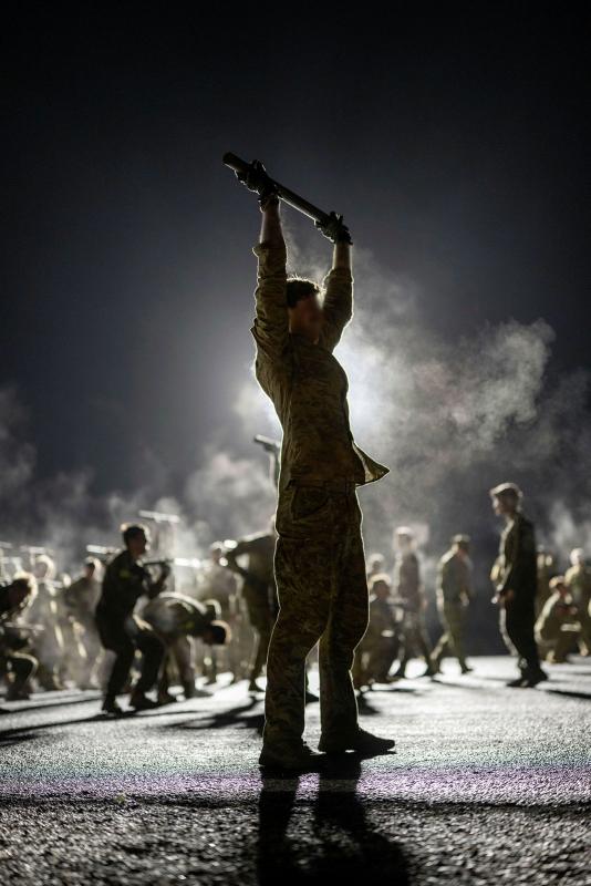 An Army man in silhouette holding up a pole above his head, with military personnel and smoke in the background.