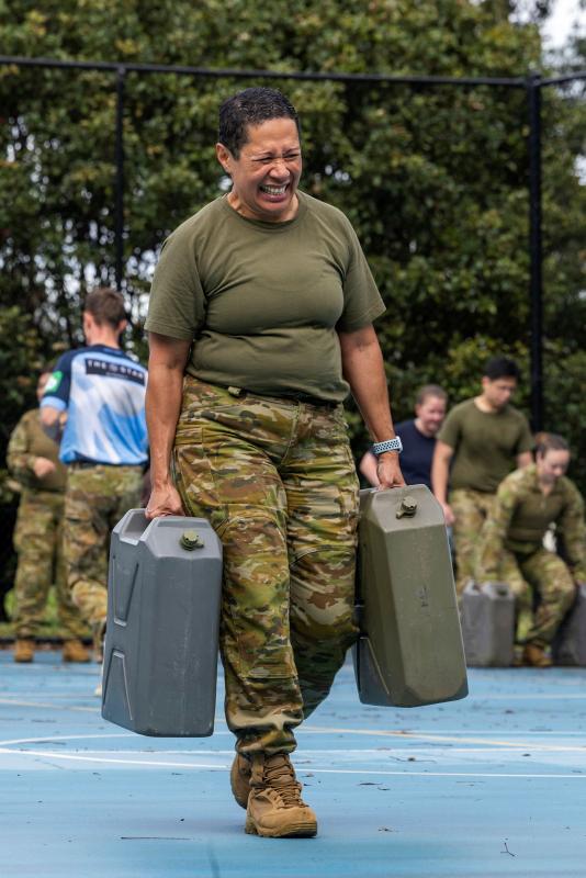A woman in military uniform struggles to carry two jerry cans.