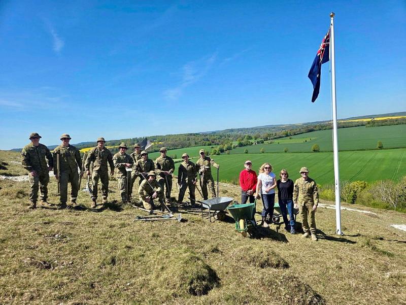 A group of people, many in uniform, stand on a hillside with gardening tools and wheelbarrows.