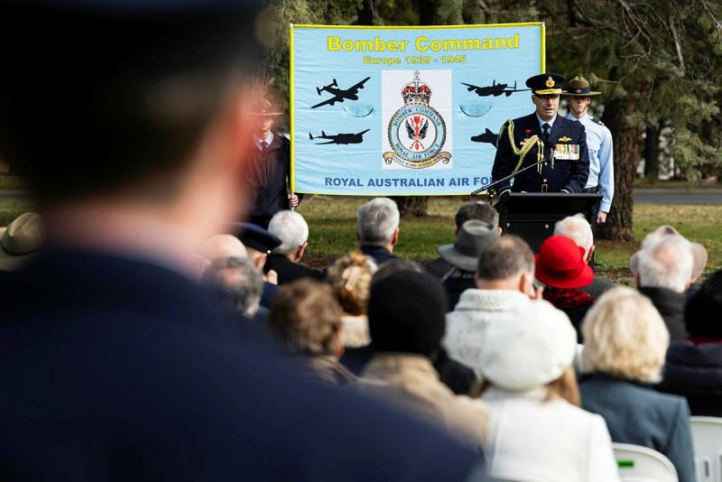 A man in military uniform addresses a crowd, with a banner for Bomber Command behind him.