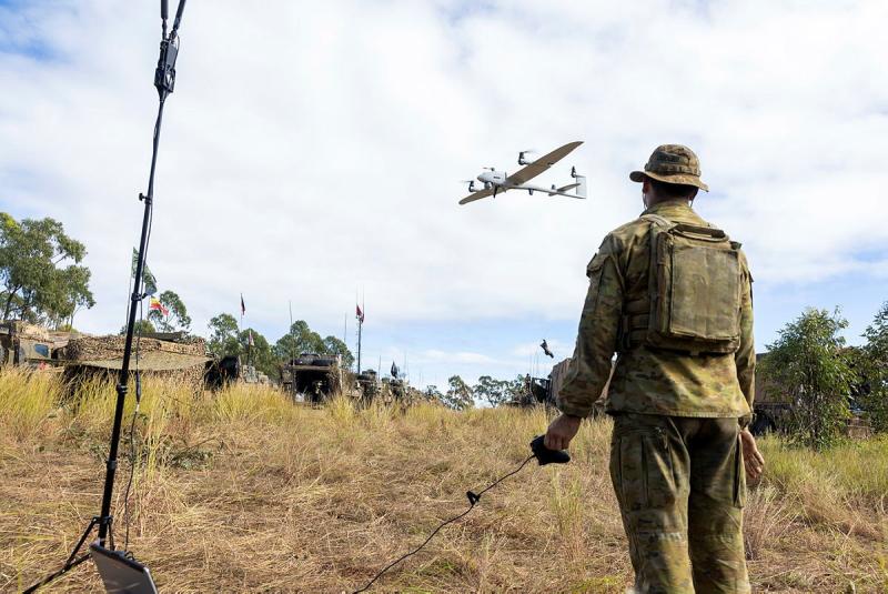 A small aircraft flies above a man in Army uniform who is holding a remote control.