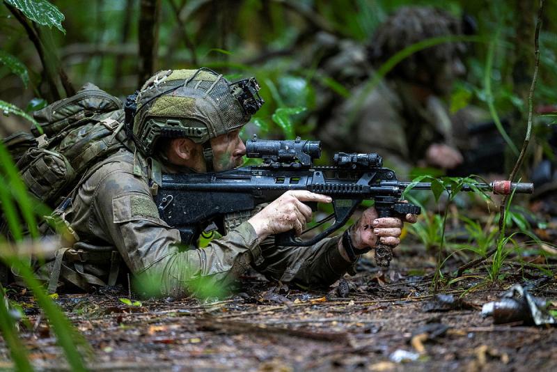 A man in a camouflage uniform and helmet lies on his stomach in the jungle aiming a rifle.