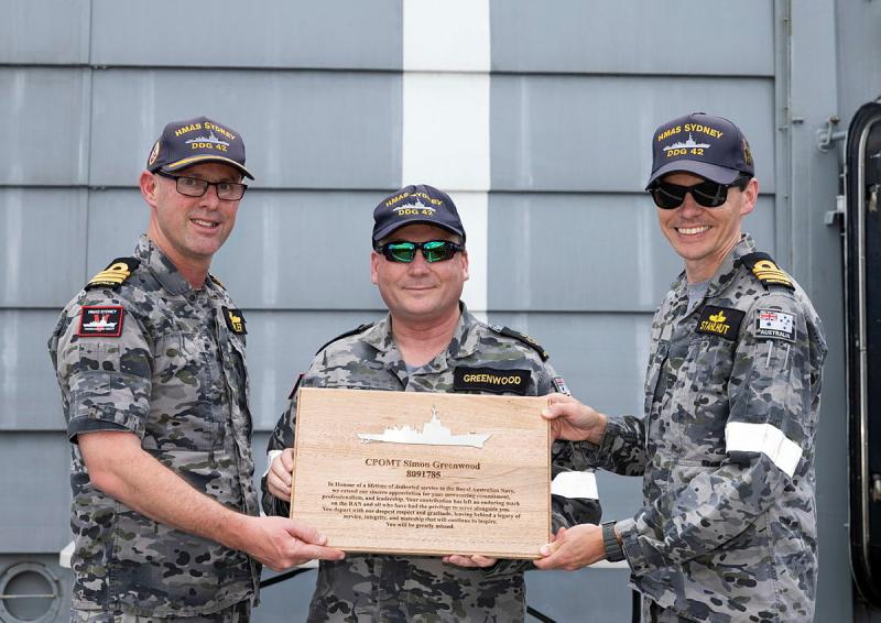 Two Navy members present another, standing in between them, with a wooden plaque.