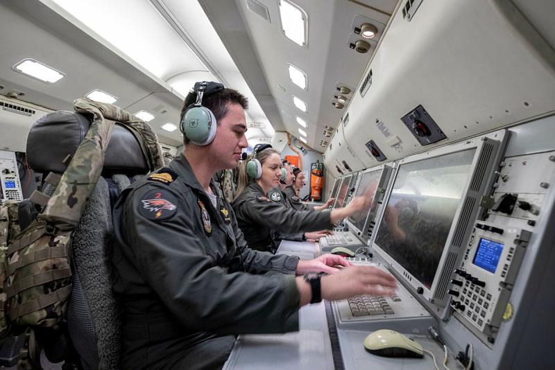 People in flight suits and headsets sit at computer workstations inside an aircraft.