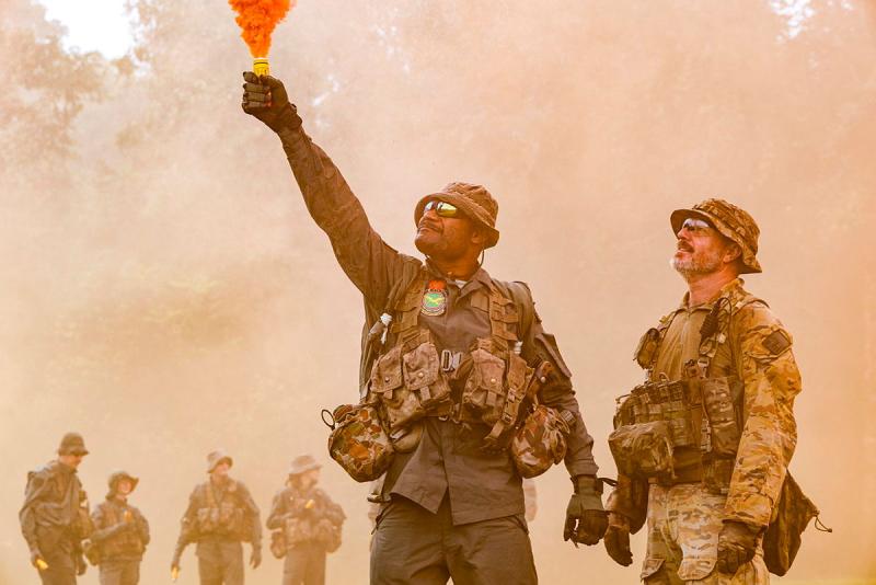 Two military personnel light an orange flare outside.