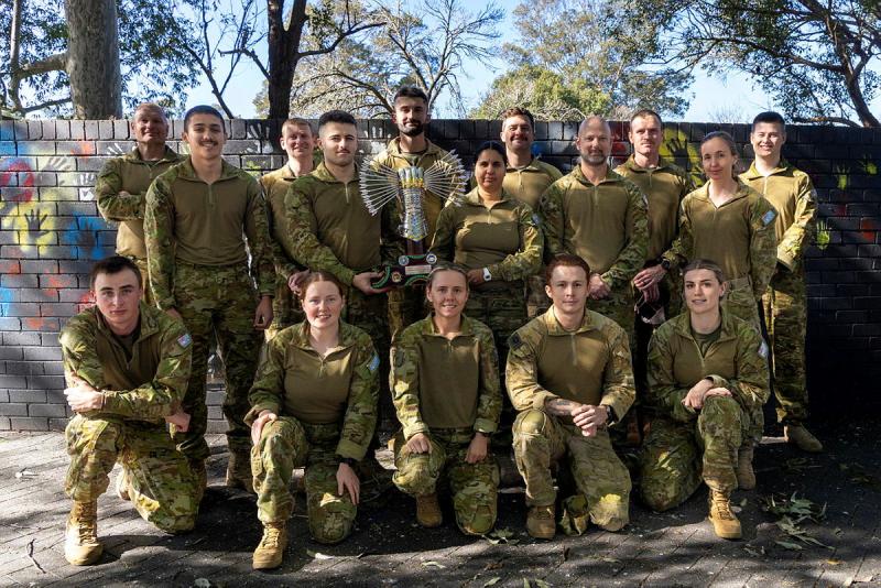 A group of military personnel stand or kneel with a shared trophy.