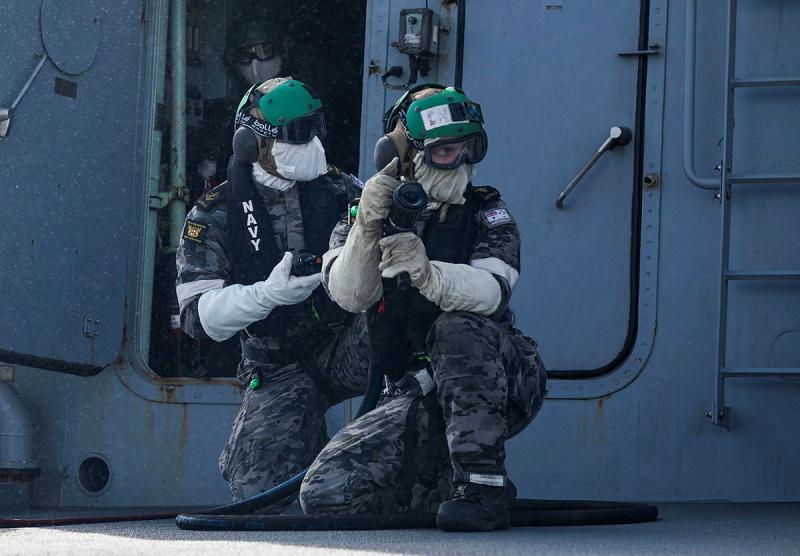 Two people in uniforms, goggles and masks kneel while holding a hose.