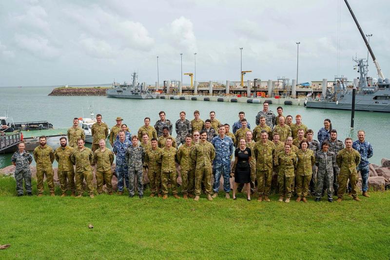 A large group of military personnel gather for a photo in front on water and ships.