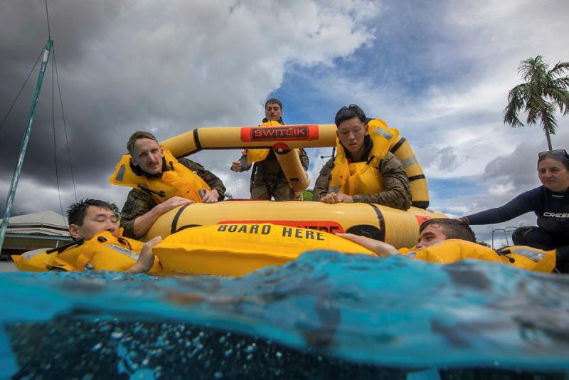 A small group of people with lifejackets on are seen in water and some on a life raft.