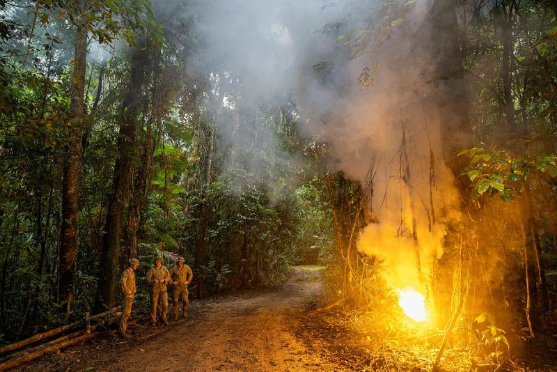 Three people in camouflage stand across a dirt track from a small fire in the middle of a jungle.