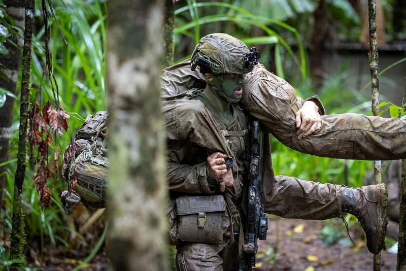 A man in camouflage and a helmet carries another person over his shoulders in a jungle.