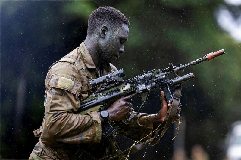 A man in camouflage uniform looks down at a rifle while standing in the rain.