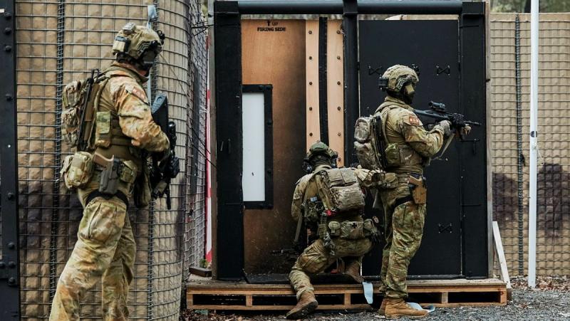 Two people in uniform stand near doors with weapons raised while another person in uniform kneels in front of the doors.
