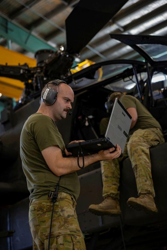 A man in uniform uses a laptop beside a helicopter while another person works next to the cockpit.