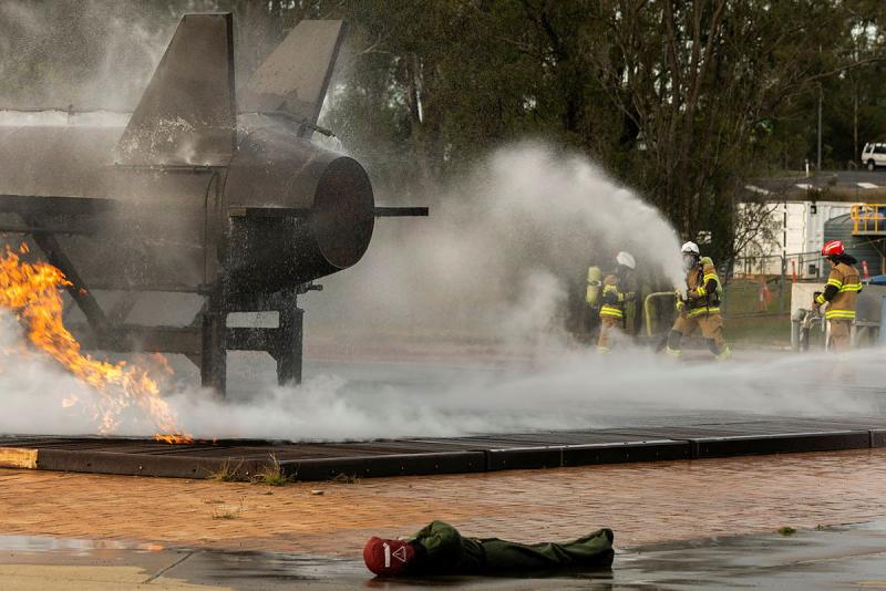 Three people in firefighting uniforms spray water at an aircraft with flames and smoke around it, while a dummy lies nearby.