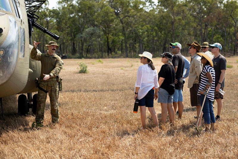 A man in uniform gestures next to a helicopter while people in casual clothes stand nearby facing the helicopter.