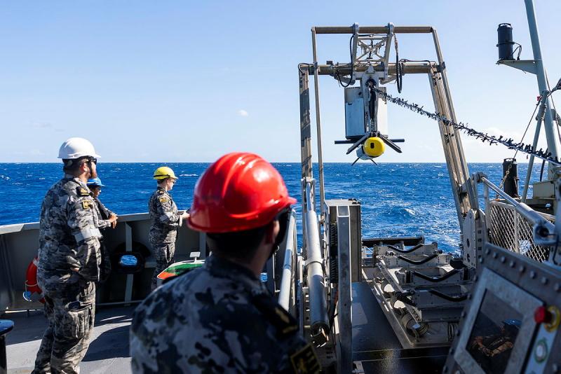 Military members aboard a ship load something on a pulley system.