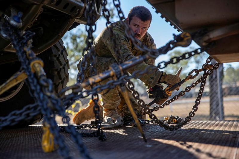 A military member makes sure the chains holding two vehicles together are secured.